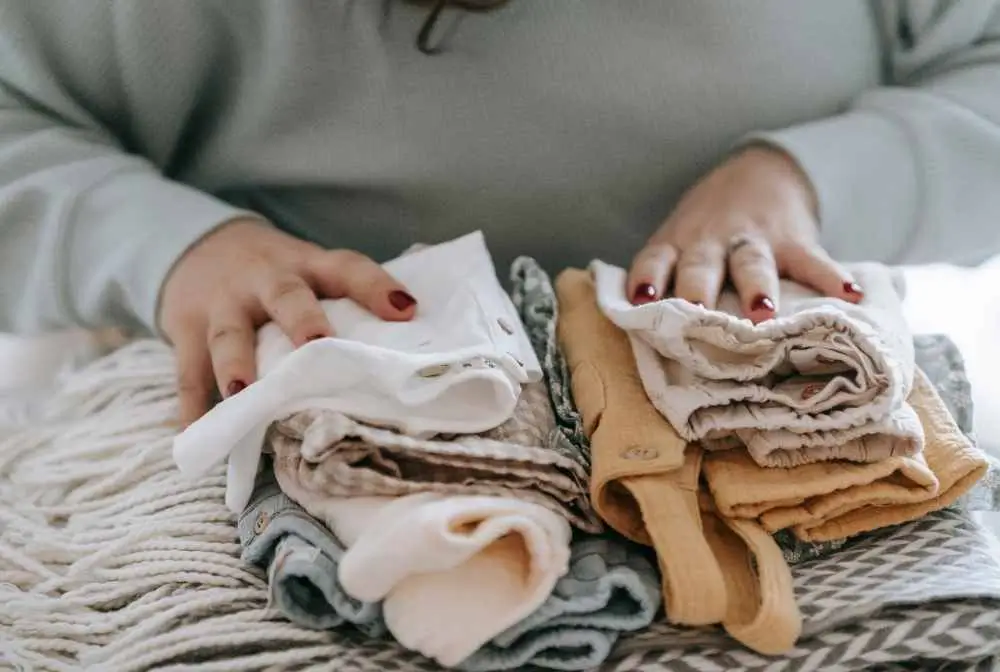 imagem de roupinhas de bebe dobradas sob móvel e mãos de uma mulher apoiadas em cada uma das pilhas