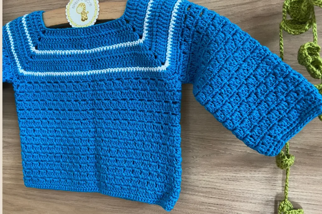 Detalhe da textura dos pontos de crochê do casaquinho para bebê azul royal, mostrando uniformidade e qualidade da confecção artesanal com etiqueta da Oficina de Crochê