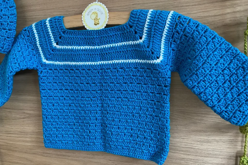 Detalhe do detalhe em fio branco na pala do casaquinho de crochê para bebê azul royal, mostrando acabamento artesanal refinado e etiqueta da marca