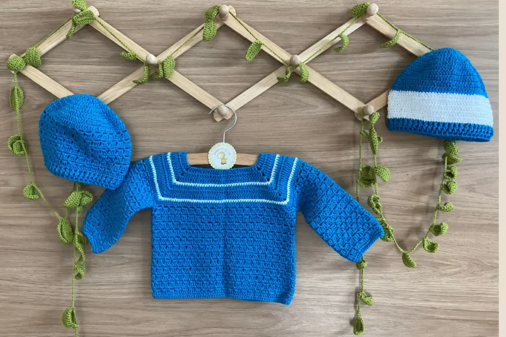 Conjunto de casaquinho e duas toucas de crochê para bebê em azul royal com detalhes brancos, dispostos sobre cabides de madeira e mesa rústica com decoração de folhas verdes