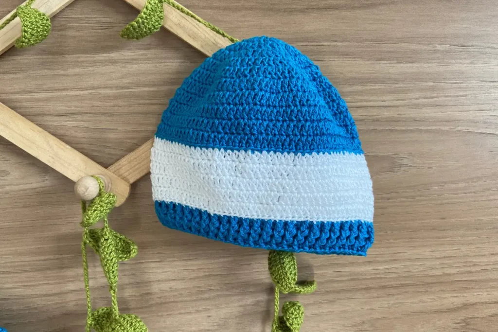 Touca de crochê para bebê com listras azuis e brancas, primeiro modelo incluído no conjunto, sobre cabides de madeira e decoração rústica