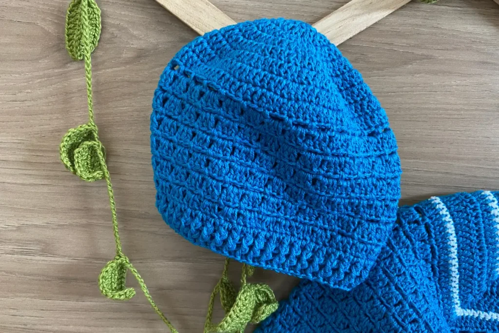 Touca de crochê para bebê em azul royal sólido com textura em pontos artesanais, segundo modelo incluído no conjunto, sobre mesa de madeira com decoração rústica